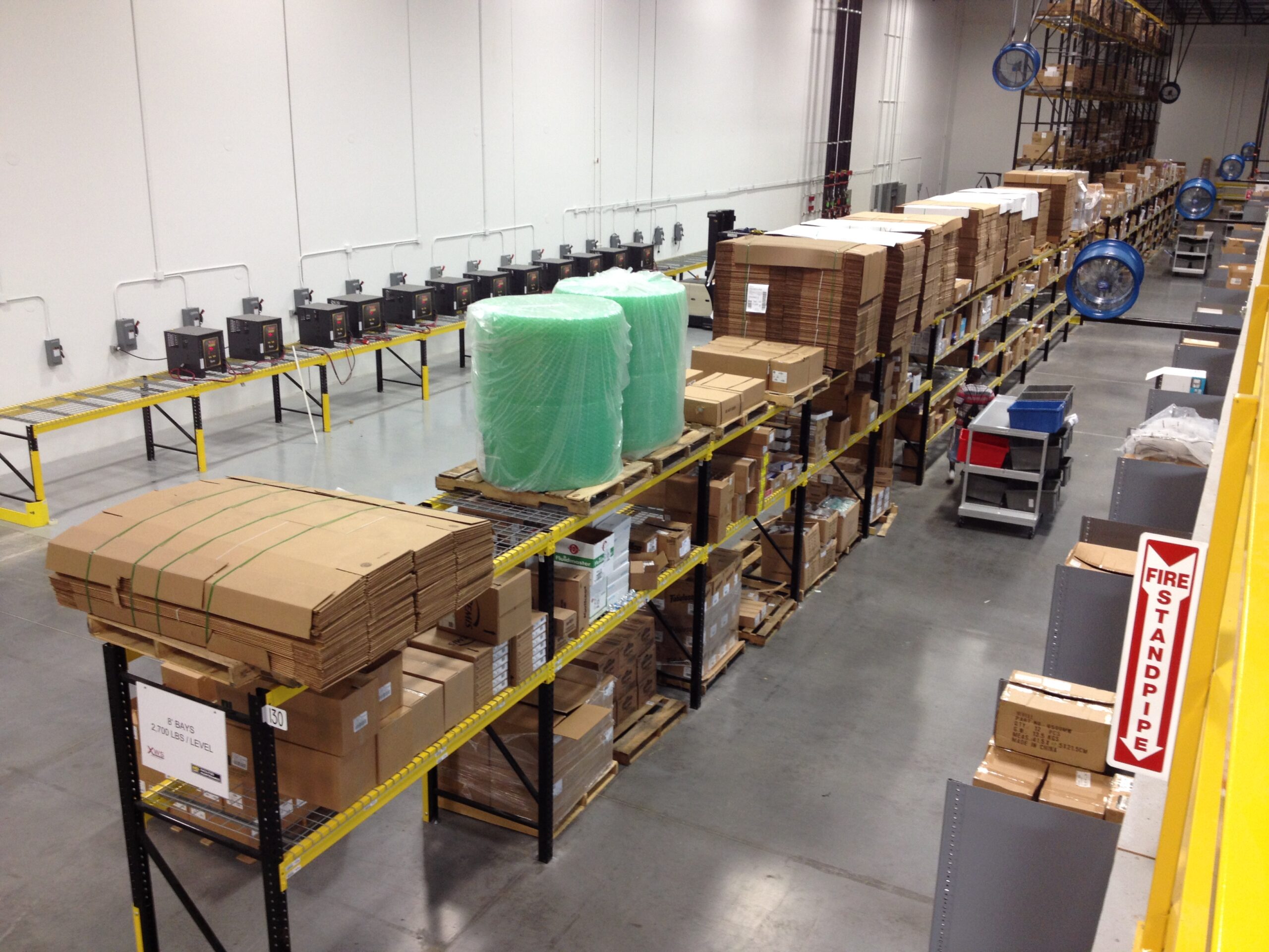 Industrial warehouse with organized shelves holding cardboard boxes, packing materials, and supplies; Material Handling Equipment is strategically placed throughout, with a fire standpipe visible in the foreground.