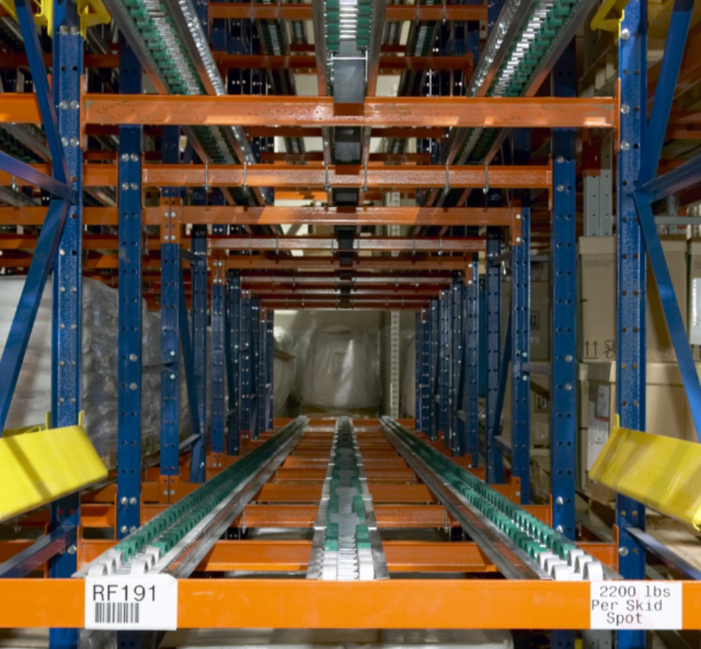 This pallet flow rack features a warehouse shelving unit with orange beams and blue supports, complete with gravity roller tracks for efficient pallet storage. A sign indicates a capacity of "2200 lbs Per Skid Spot.