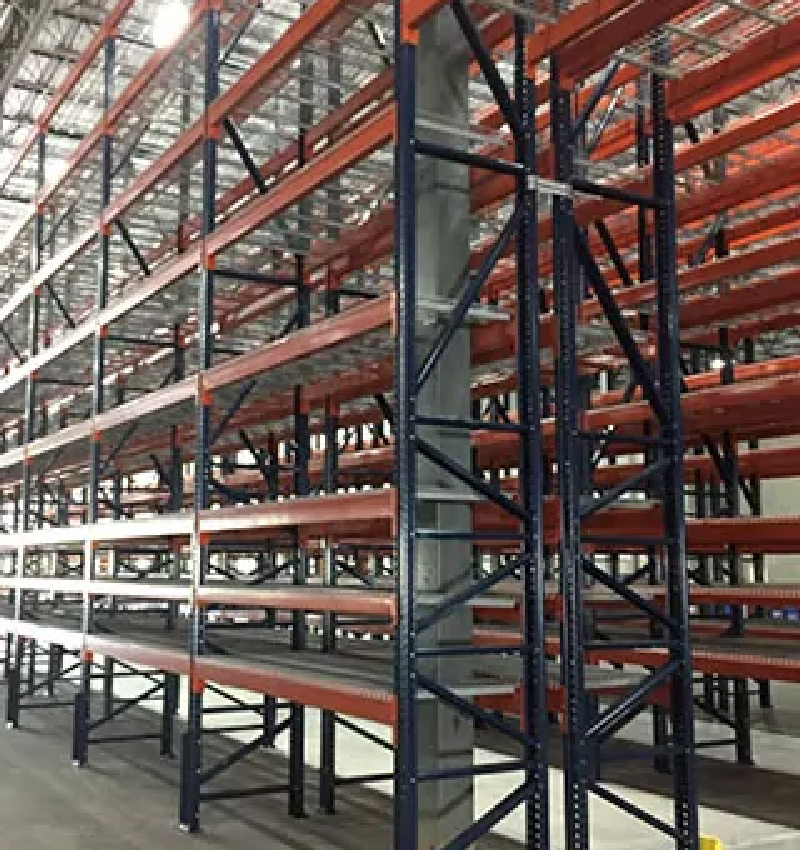 Empty industrial warehouse shelving units made of metal, commonly used as material handling equipment, are arranged in rows under a high ceiling with exposed beams and bright overhead lighting.