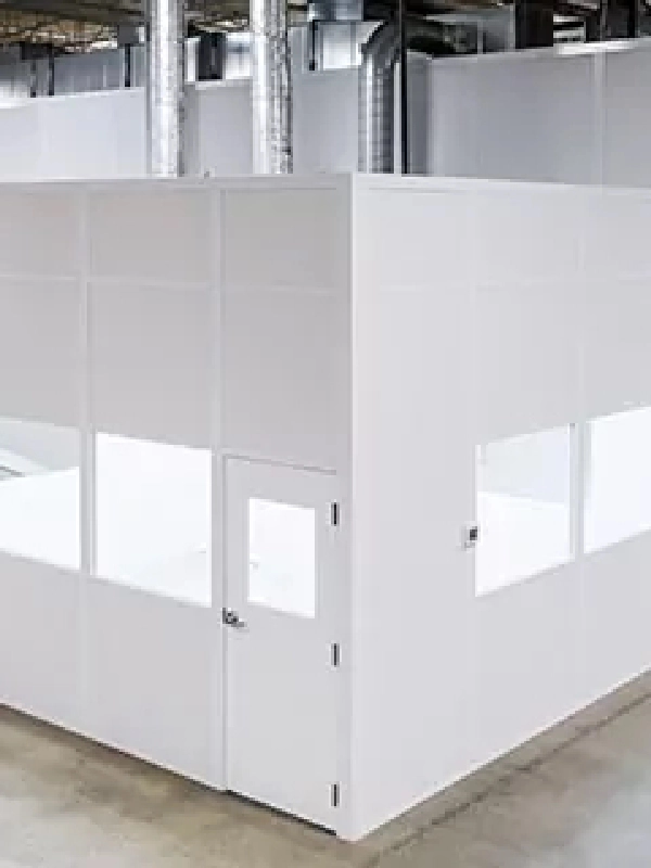 A white modular office, part of innovative modular buildings, with windows and a closed door stands inside a large industrial warehouse with visible ductwork above.