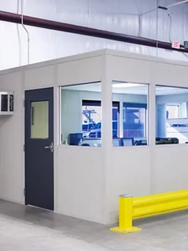 A small, enclosed modular office with windows and a black door is inside a warehouse, featuring a yellow safety barrier out front. Modular buildings like this offer flexible workspace solutions.