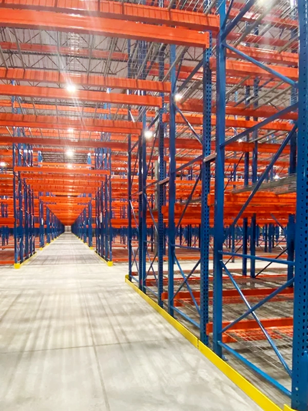 Empty industrial warehouse with tall blue and orange Double-Deep Rack shelving units, evenly spaced on a concrete floor under bright ceiling lights.