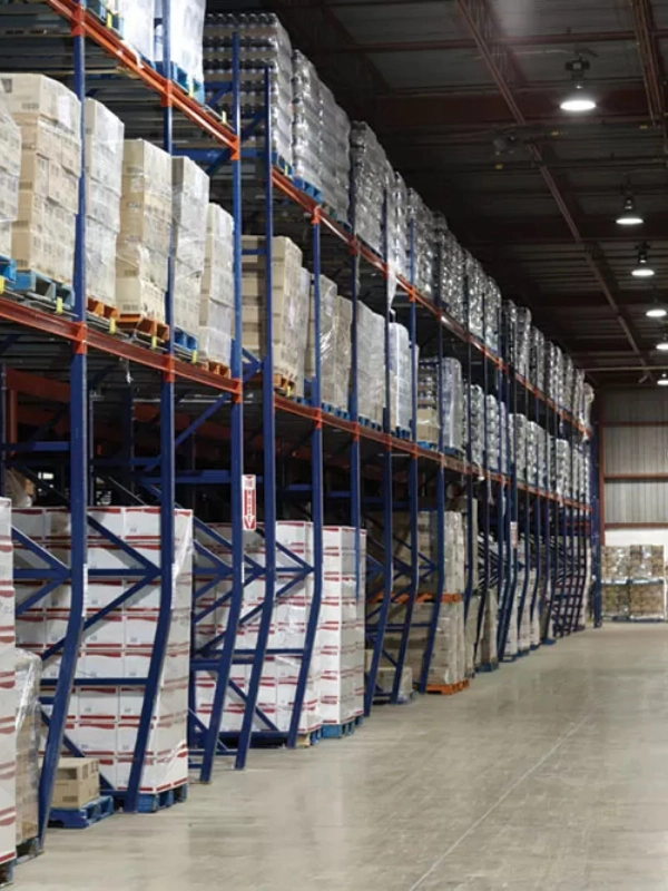 Tall industrial warehouse racks, including Drive-In Rack systems, filled with stacked pallets of boxed goods line a clean, spacious concrete floor under bright ceiling lights.