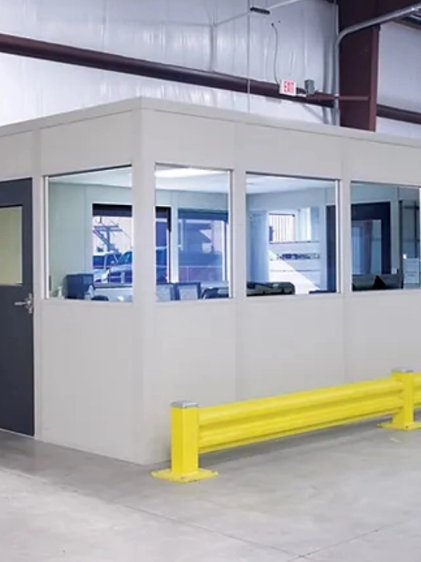 A prefabricated office with large windows and a dark door is situated inside a warehouse. Warehouse safety equipment, including a yellow safety barrier, is positioned in front of the structure.