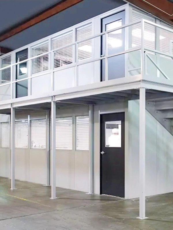Two-story modular office with white panels and black doors inside a warehouse, featuring stairs and large windows on both levels. Discover the flexibility of Modular Buildings for efficient, modern workspaces.