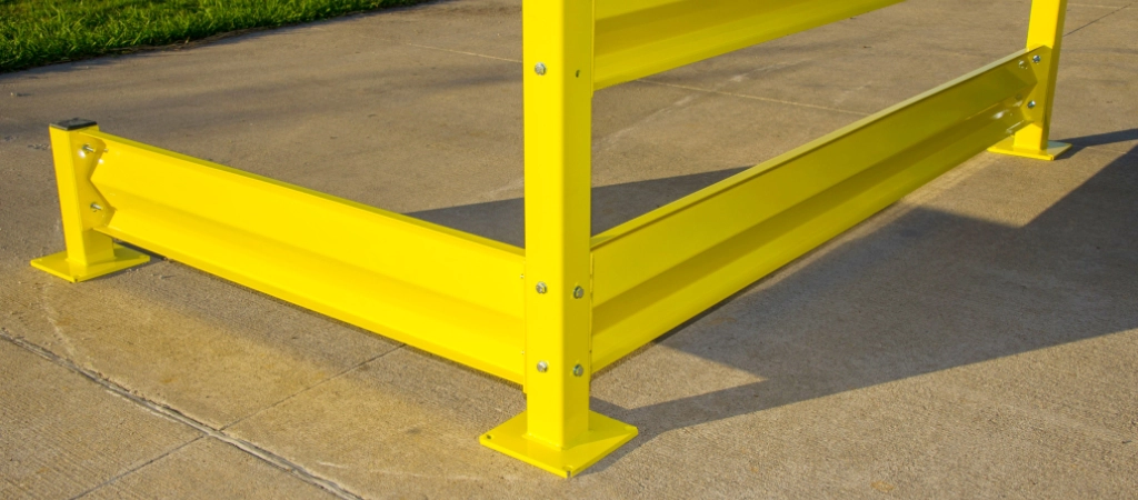 Yellow metal safety barrier with bolted base plates installed on a concrete surface outdoors, next to green grass. Ideal as warehouse safety equipment, it provides strong protection for workspaces and outdoor areas.