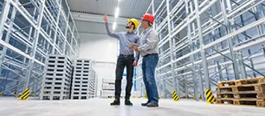 Two men wearing safety helmets stand in an empty warehouse, discussing or inspecting the storage racks and facility during a turnkey warehouse project.