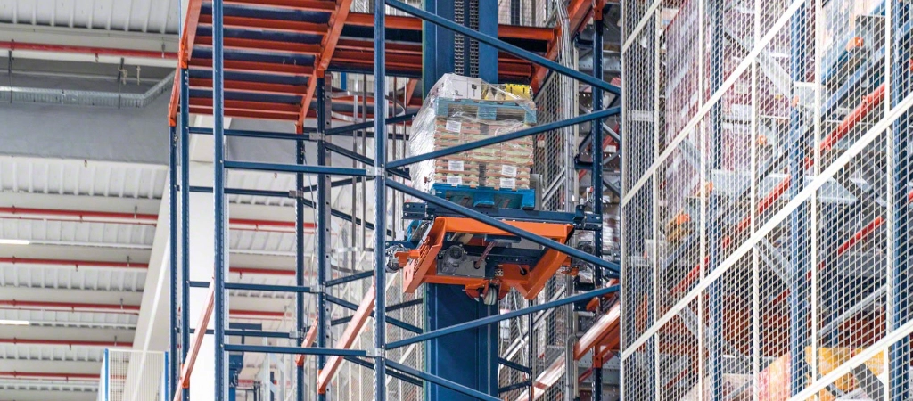 Automated warehouse system lifts a pallet of goods to a high storage shelf using a vertical conveyor, metal support structure, and integrated warehouse safety equipment.
