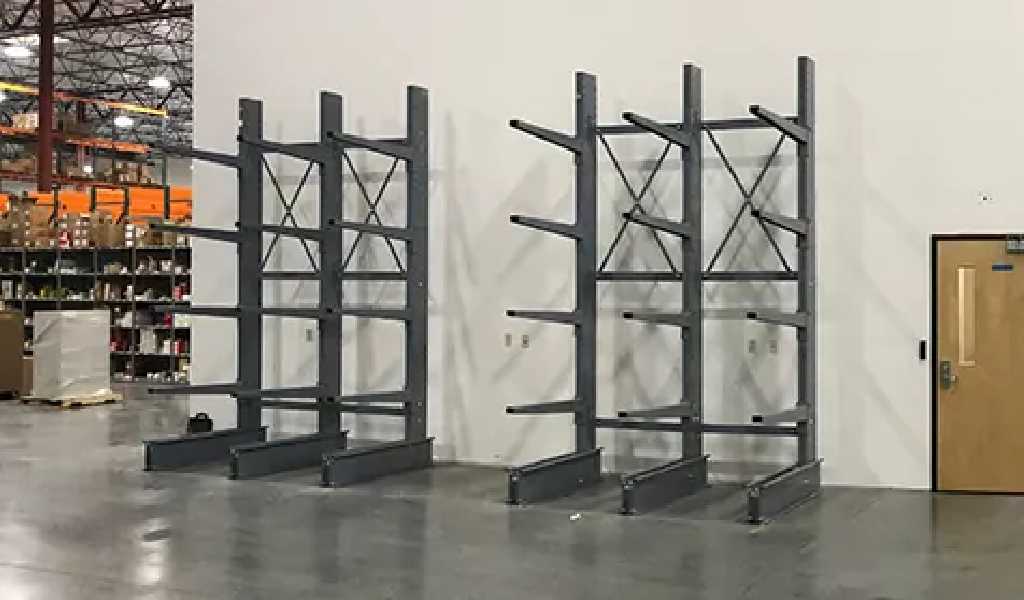 Empty metal warehouse shelving units, including a cantilever rack, are positioned against a white wall on a polished concrete floor, with a door to the right and storage shelves visible in the background.