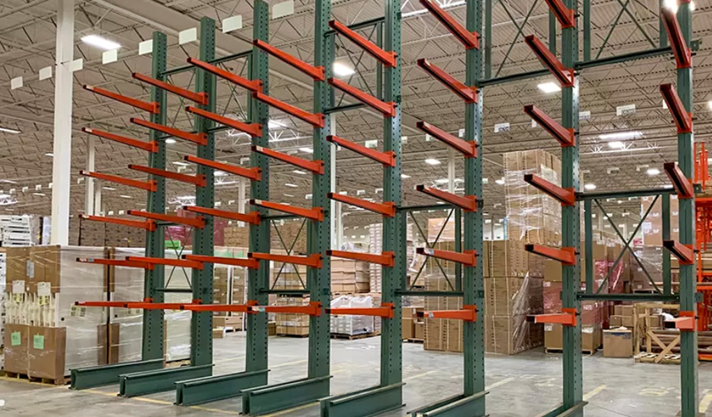Empty green metal Cantilever Racks with orange arms are installed in a warehouse, with boxes and pallets stacked in the background.