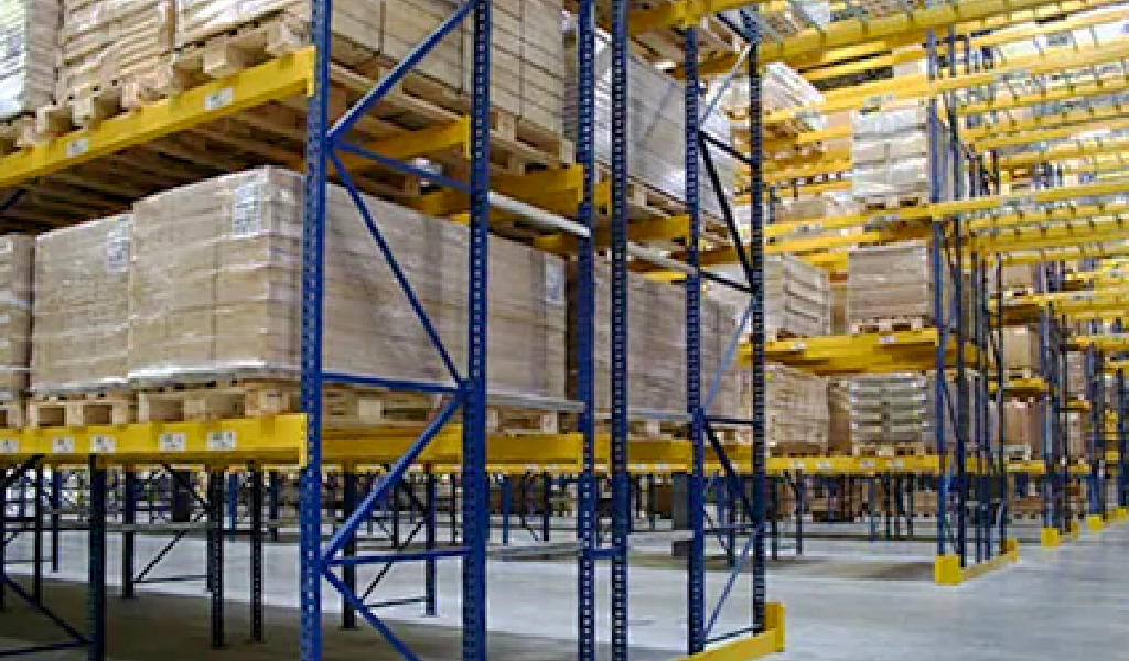 Pallets stacked on tall double-deep racks in a warehouse with a concrete floor and yellow safety barriers.