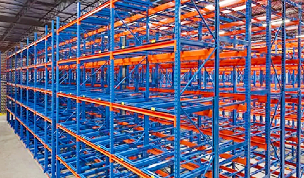 Empty industrial warehouse shelving units with blue frames and orange beams, arranged in multiple rows, resemble push-back rack systems when viewed from an angle inside the spacious warehouse.