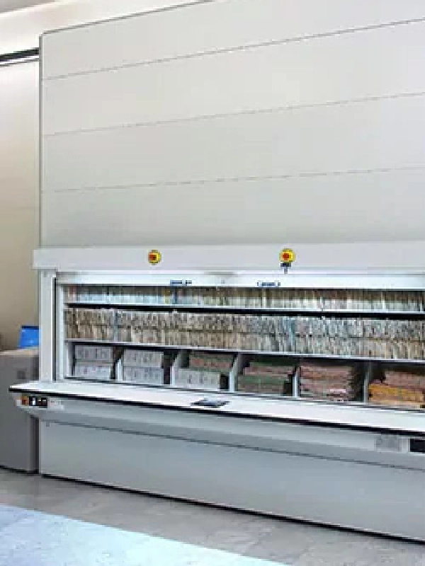 Automated vertical filing cabinet with shelves containing organized paper files and folders in an office setting, featuring efficient Vertical Lift Modules for streamlined storage and retrieval.