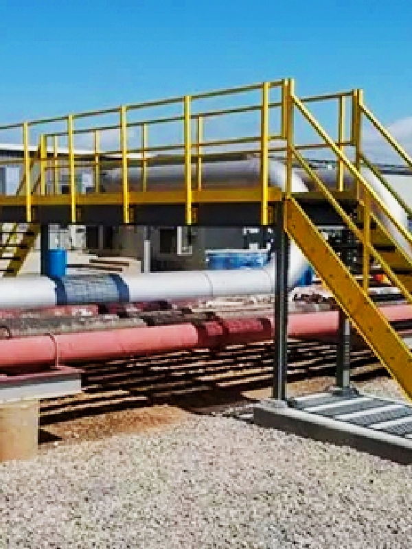 A yellow metal pedestrian bridge crosses over several large industrial pipelines at an outdoor facility, connecting steel platforms and mezzanines for safe and efficient access.