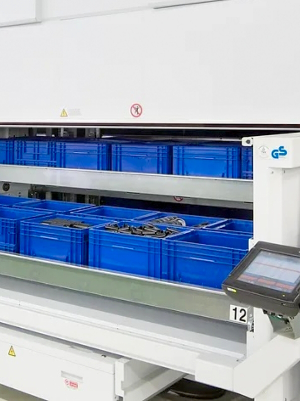 Automated storage system with blue bins on metal shelves and a touchscreen control panel in an industrial setting, featuring advanced Vertical Lift Modules for efficient space utilization.