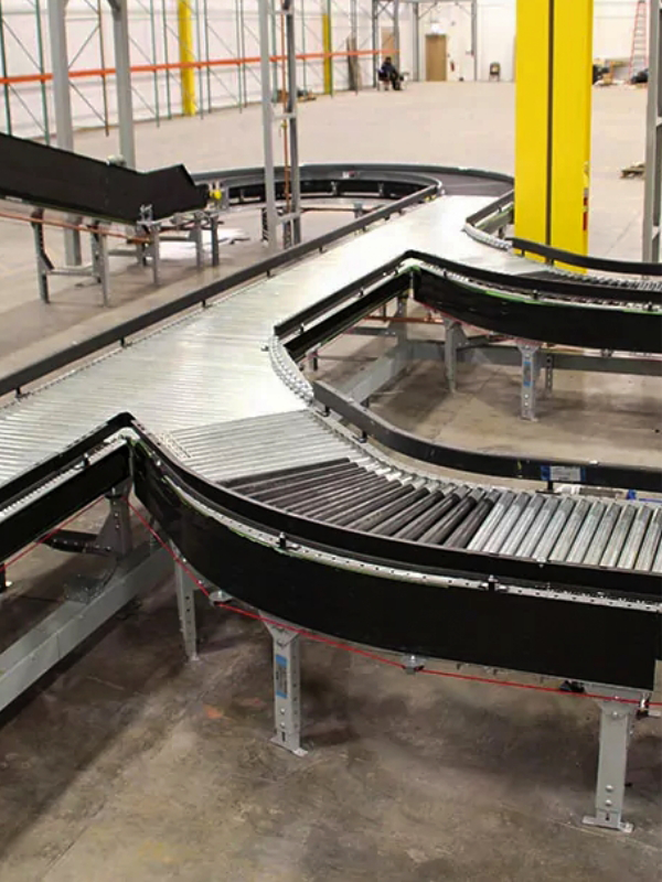 Steel roller conveyor systems with black side guards curve through an industrial warehouse featuring yellow support columns and concrete flooring.