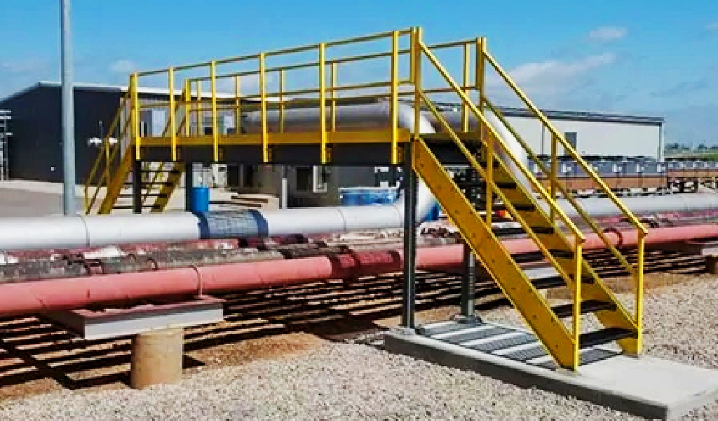 A yellow metal pedestrian bridge with stairs crosses over several large industrial pipelines at an outdoor facility, showcasing the functionality of steel platforms & mezzanines.