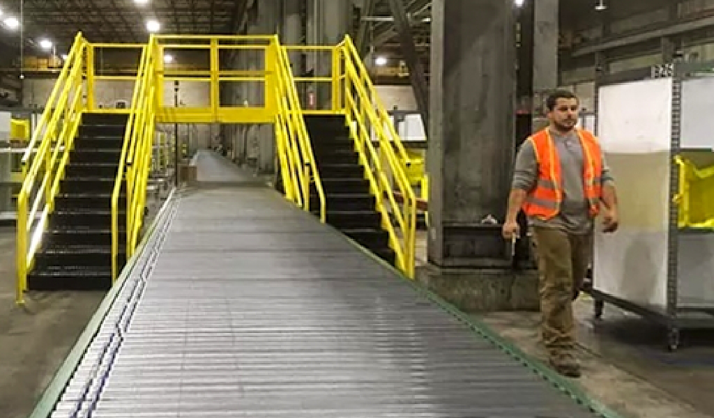 A worker in a reflective vest stands next to a conveyor belt and yellow-railed stairs, with Steel Platforms & Mezzanines visible inside an industrial warehouse.