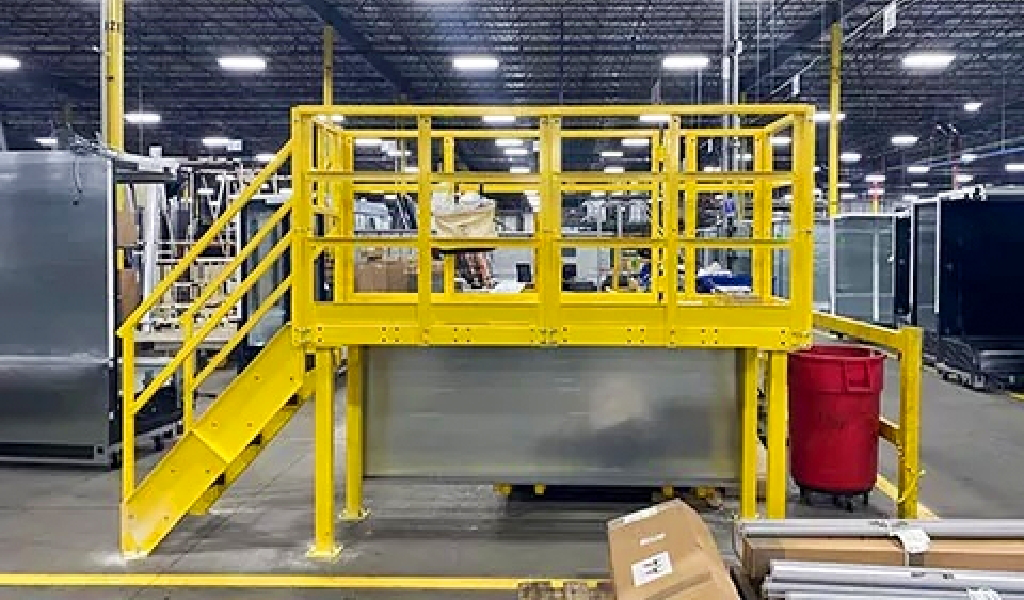 A yellow steel platform with stairs and railings, part of Steel Platforms & Mezzanines solutions, is inside an industrial warehouse; various equipment and a red trash bin are nearby.