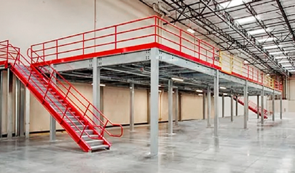 Industrial warehouse interior featuring steel mezzanine platforms, bold red railings, and dual staircases on either side; concrete floor and high ceiling with exposed beams.