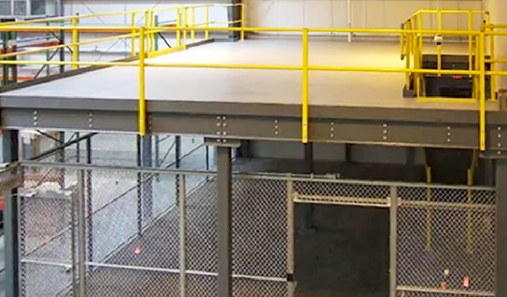 A steel mezzanine with yellow railings is built above a fenced area inside an industrial warehouse, showcasing the versatility of Steel Platforms & Mezzanines.