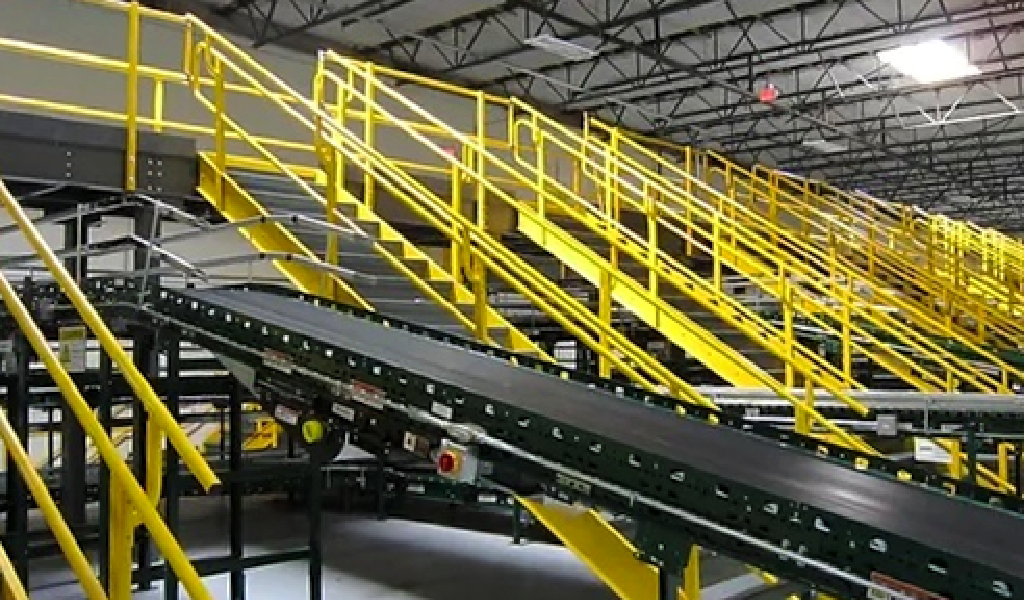 A black conveyor belt runs diagonally in a warehouse, surrounded by yellow metal railings, staircases, and steel platforms & mezzanines beneath an exposed ceiling.
